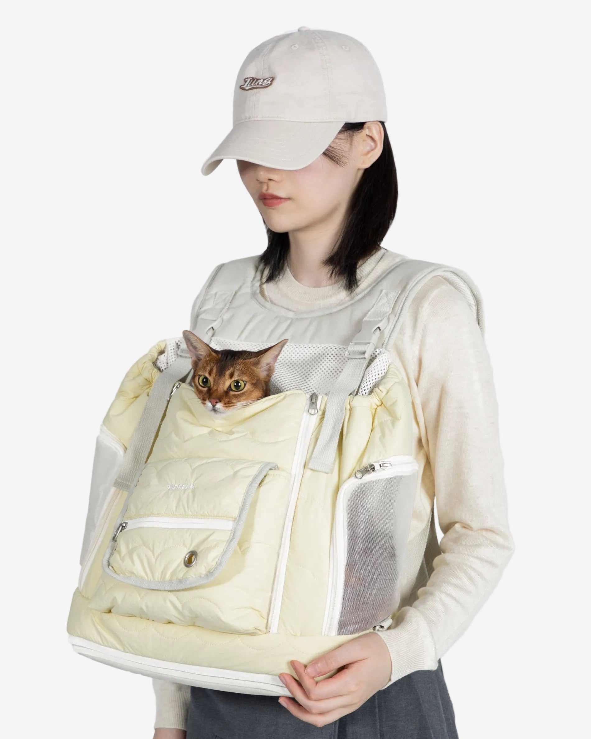 Person wearing a light-colored pet carrier with a cat peeking out, on a plain background