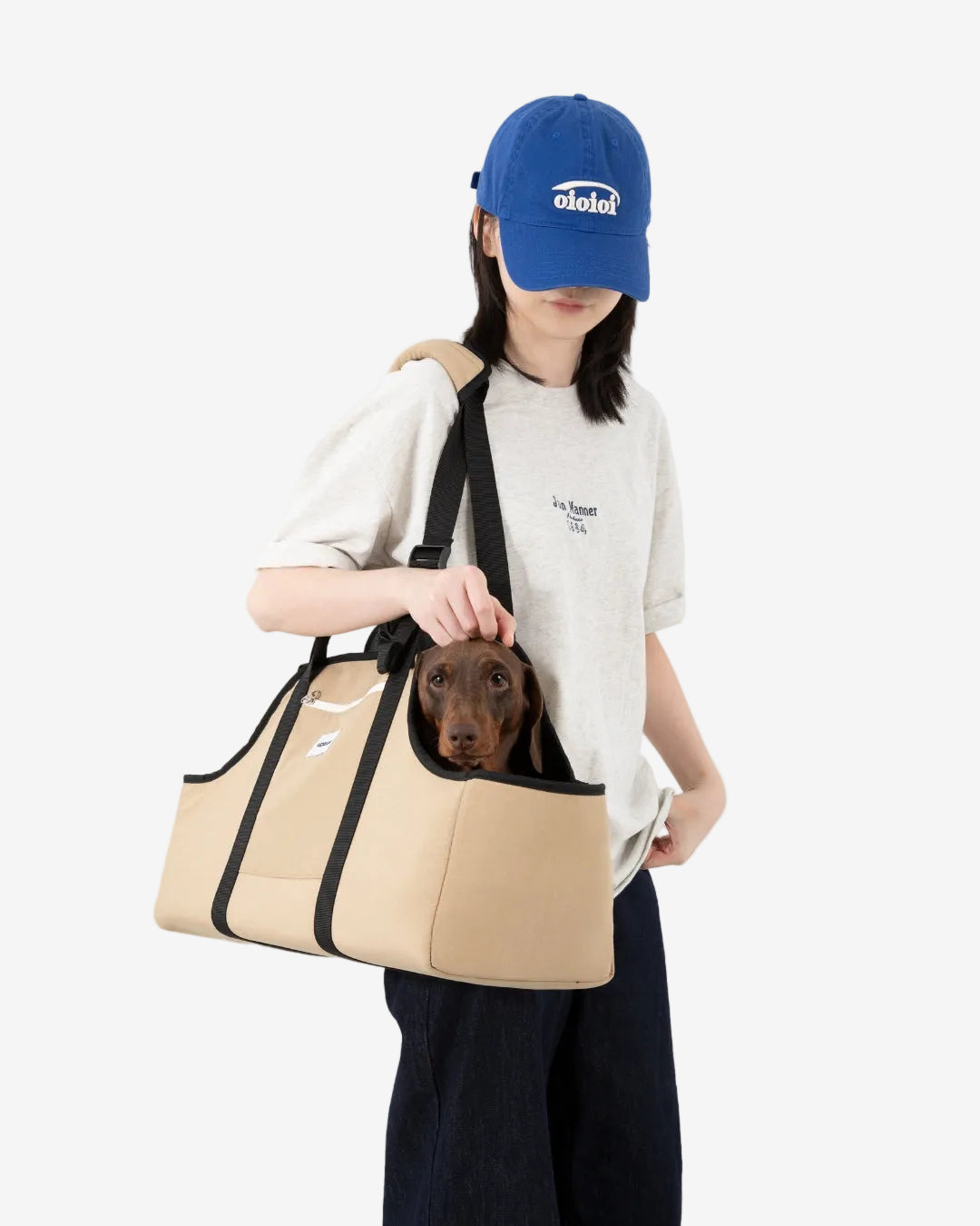 Person holding a beige Dachshund dog carrier with a dog inside, wearing a blue cap and white shirt.