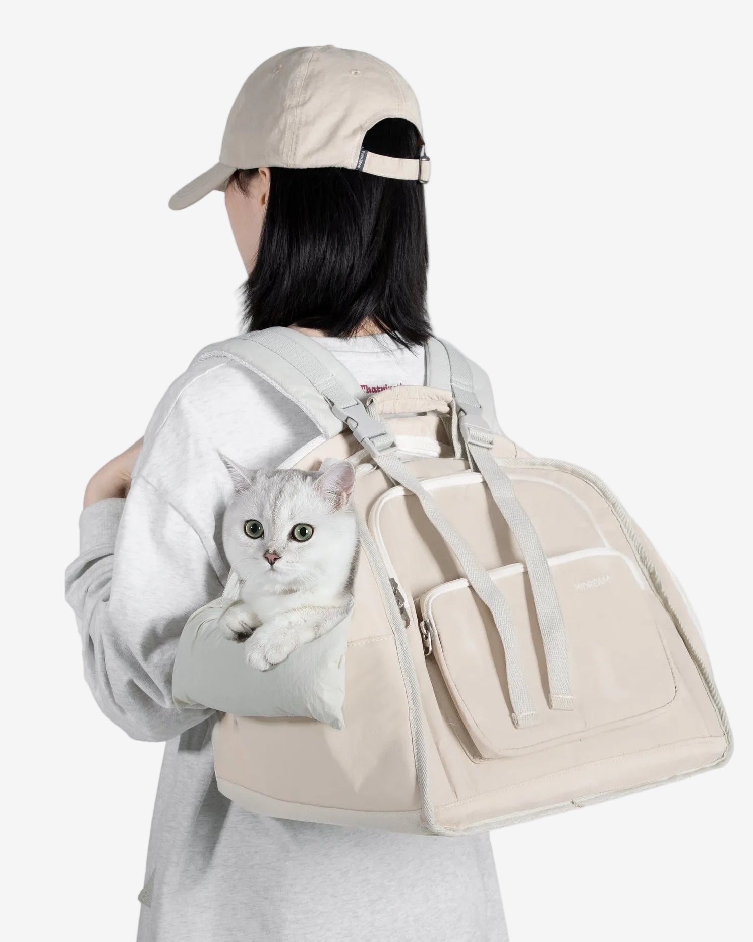 Person holding a white cat in a beige backpack against a light gray background